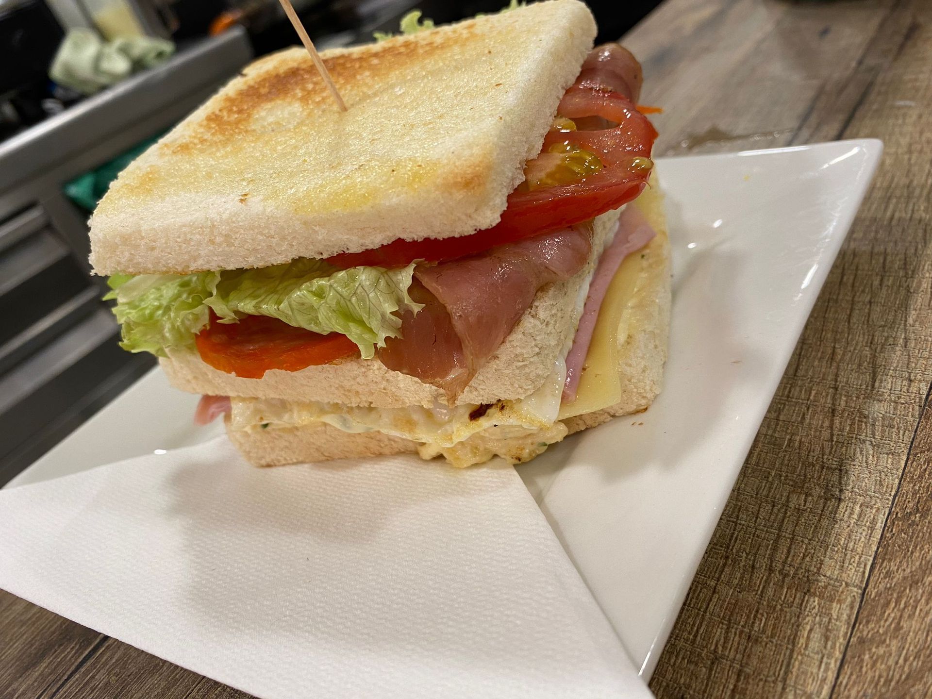 Sándwich en plato blanco con jamón, tomate, lechuga y queso, sujetos con un palillo.