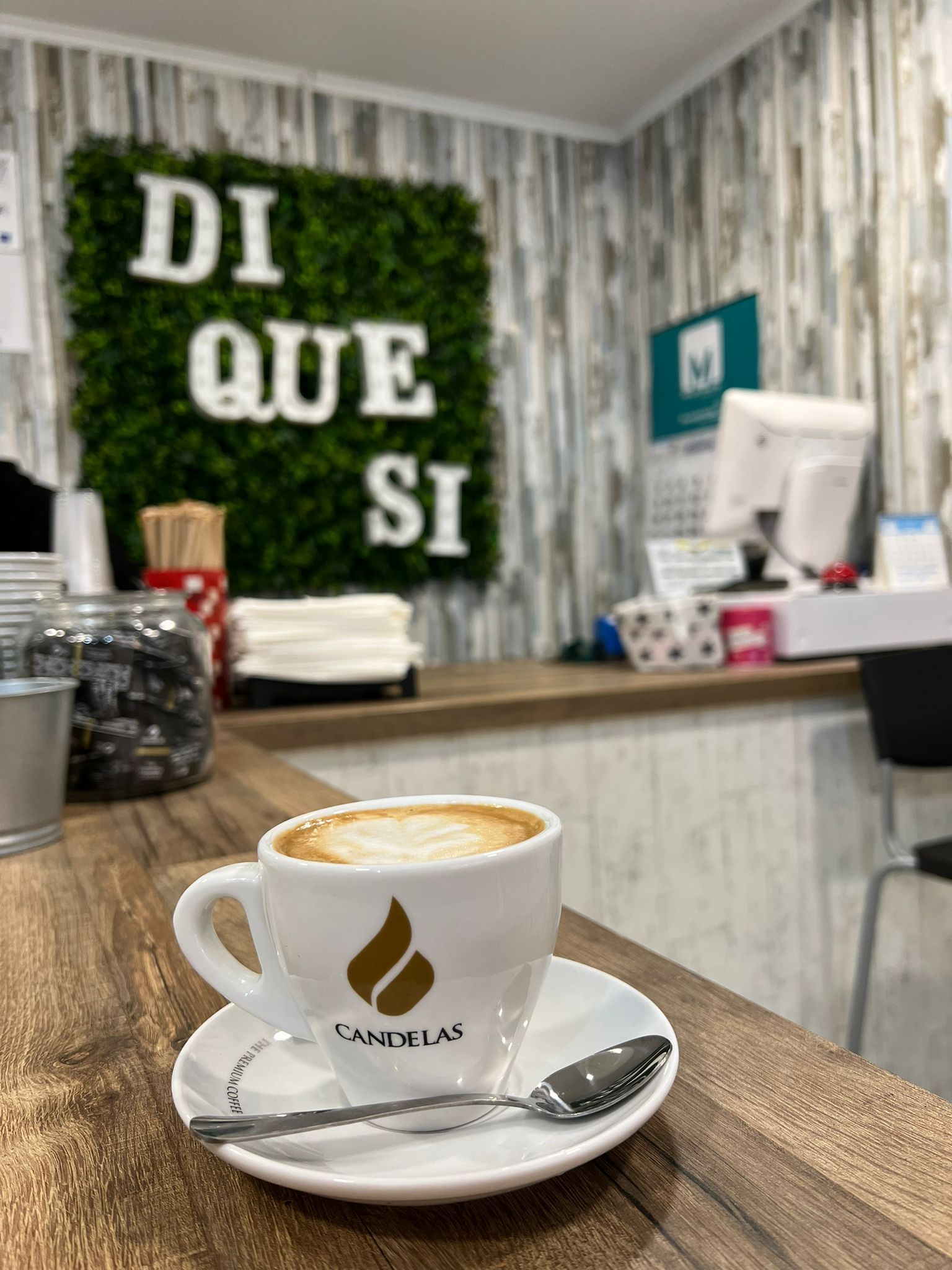 Capuchino en una taza blanca sobre un platillo, sobre un mostrador de madera con el cartel 
