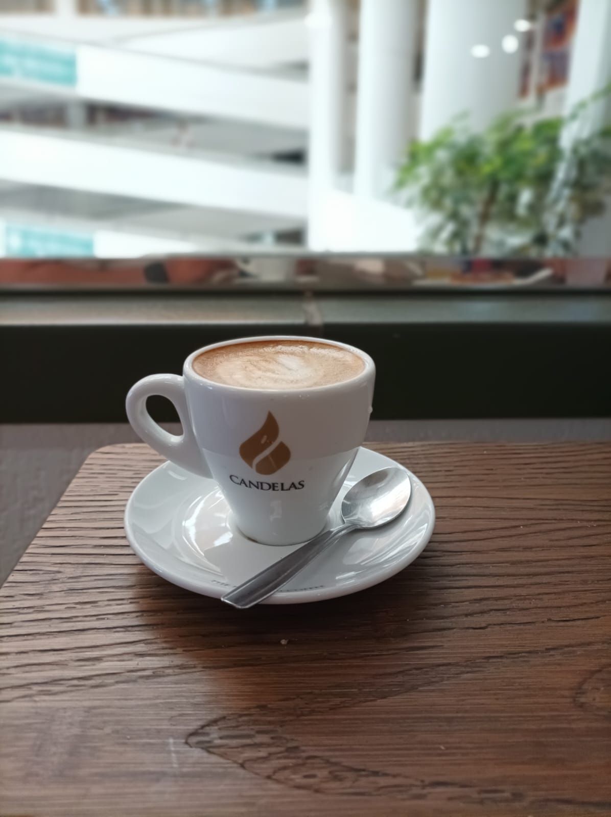 Una taza de capuchino con una cuchara en un platillo reposa sobre una mesa de madera cerca de una ventana.