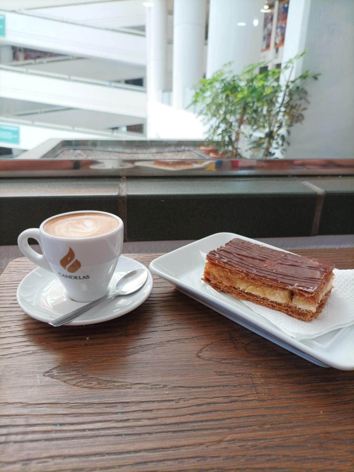Taza de café y pastel sobre una mesa cerca de una ventana, con un interior borroso y una planta al fondo.
