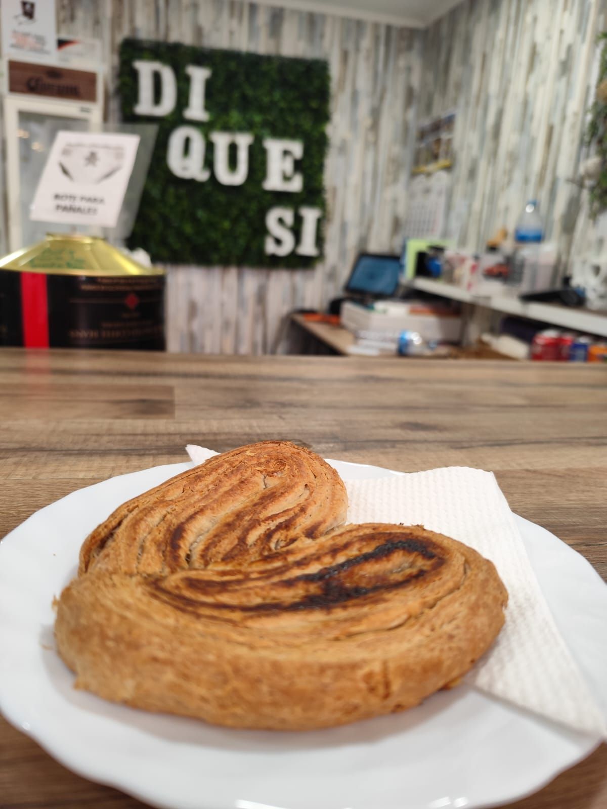 Un pastel en un plato blanco reposa sobre un mostrador de madera frente a una tienda con un cartel que dice 