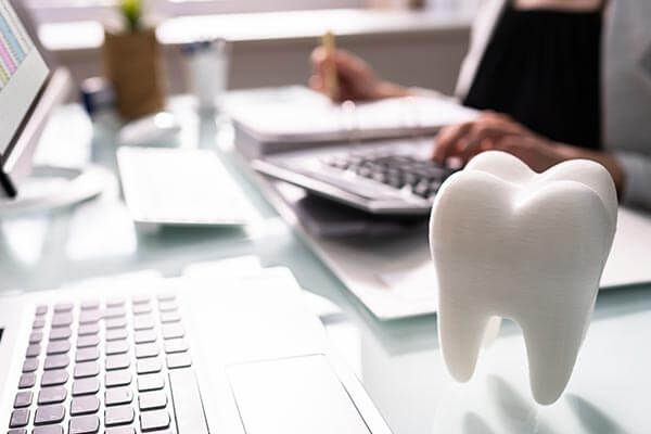 White ceramic tooth in focus on a desk with a laptop and person working on finances.