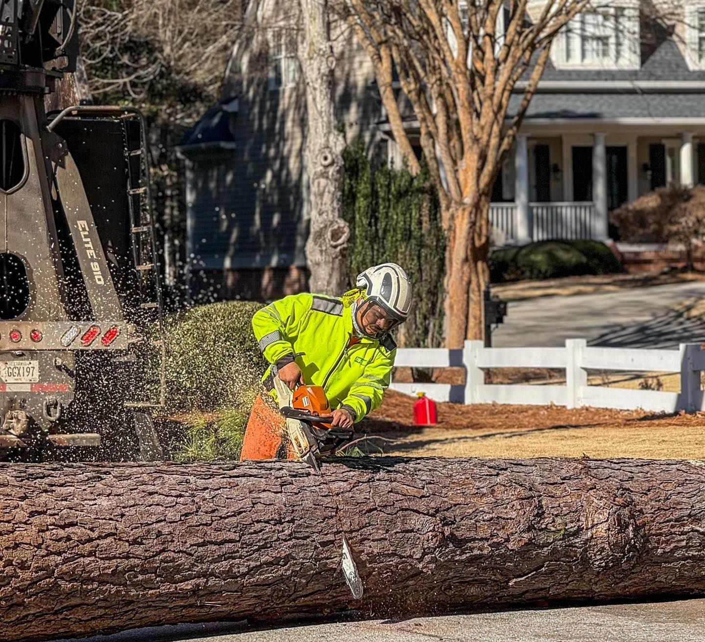 A worker in a high-visibility yellow jacket and hard hat uses a chainsaw to cut a large log outdoors near a house.