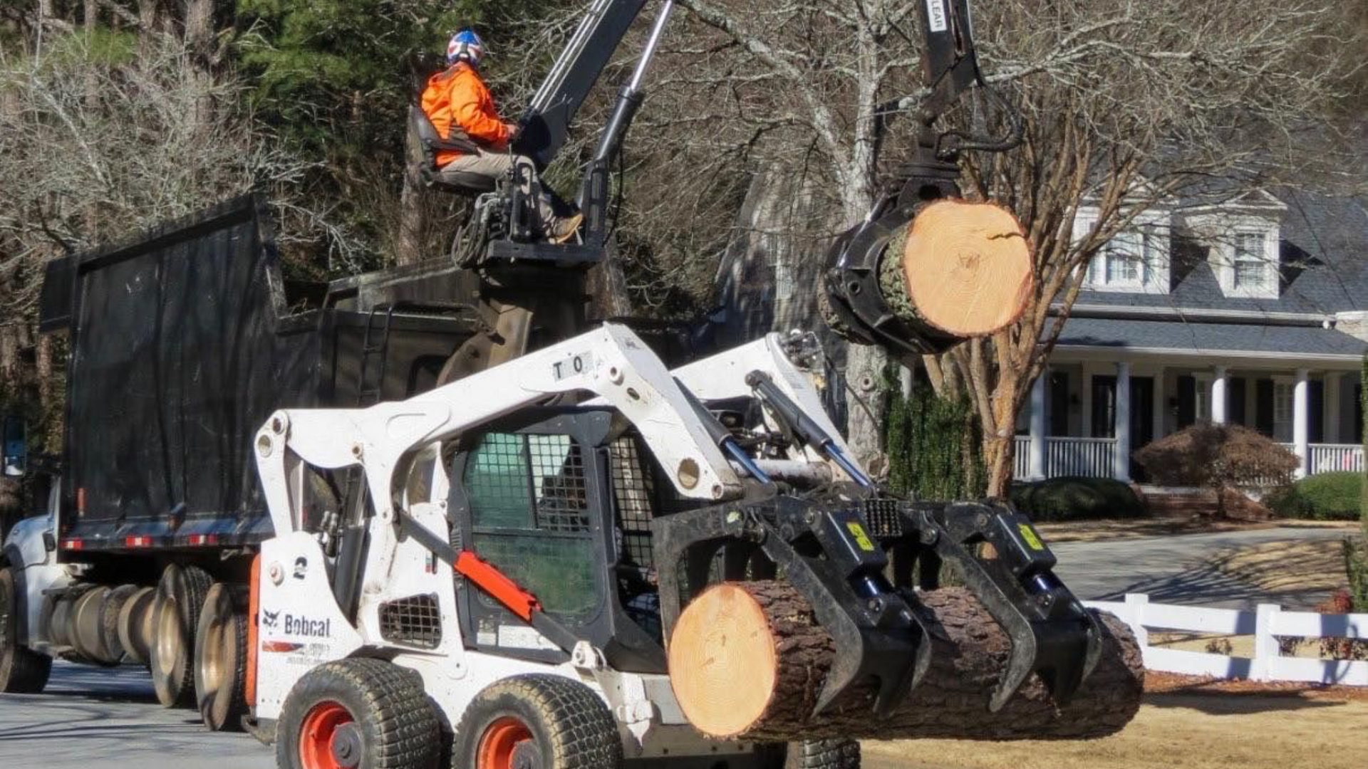 A worker operates a crane on a truck, while a white skid steer loader grips logs in a residential neighborhood.
