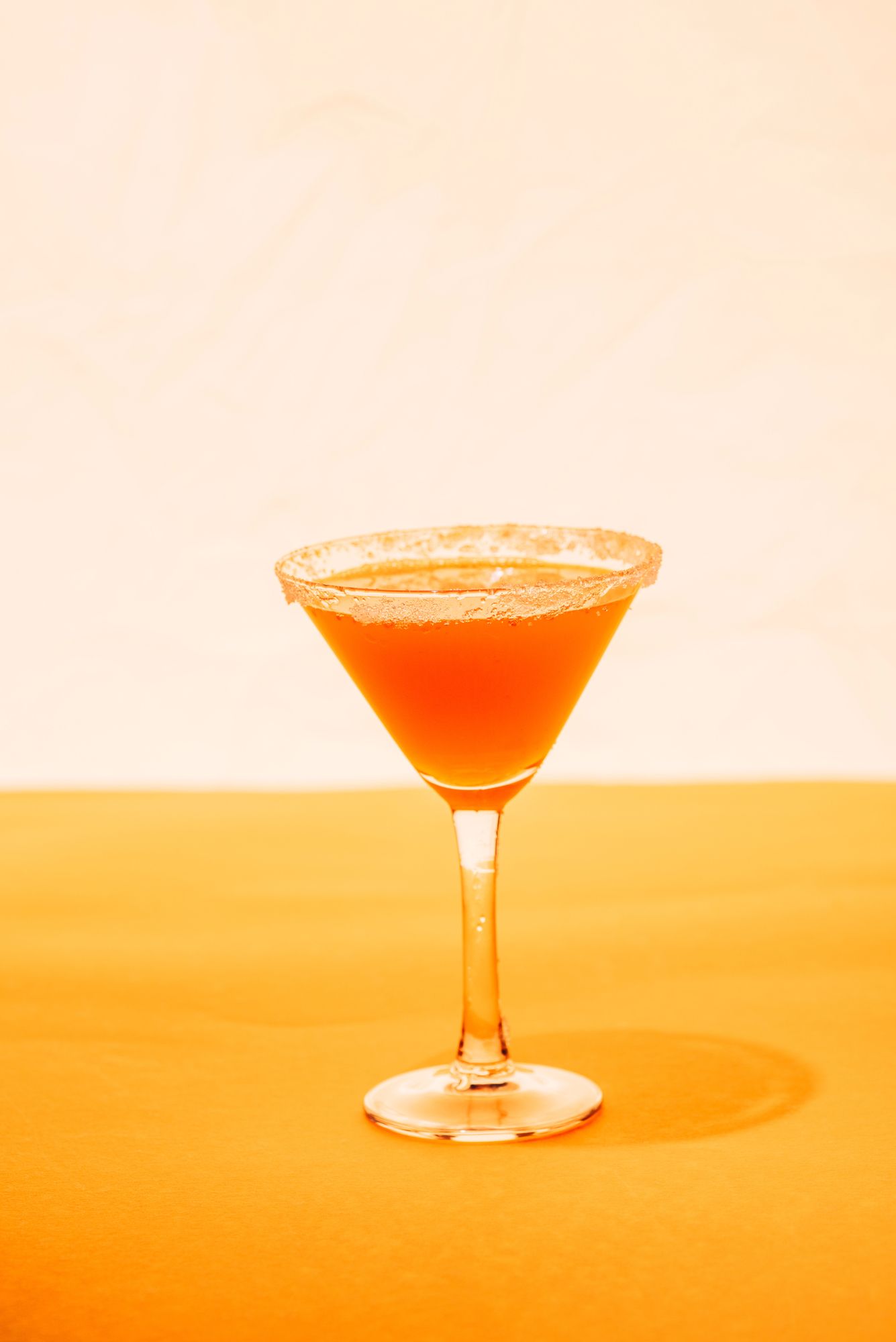 Cocktail in un bicchiere da martini con bordo zuccherato, adagiato su una superficie giallo brillante, sfondo bianco sporco.