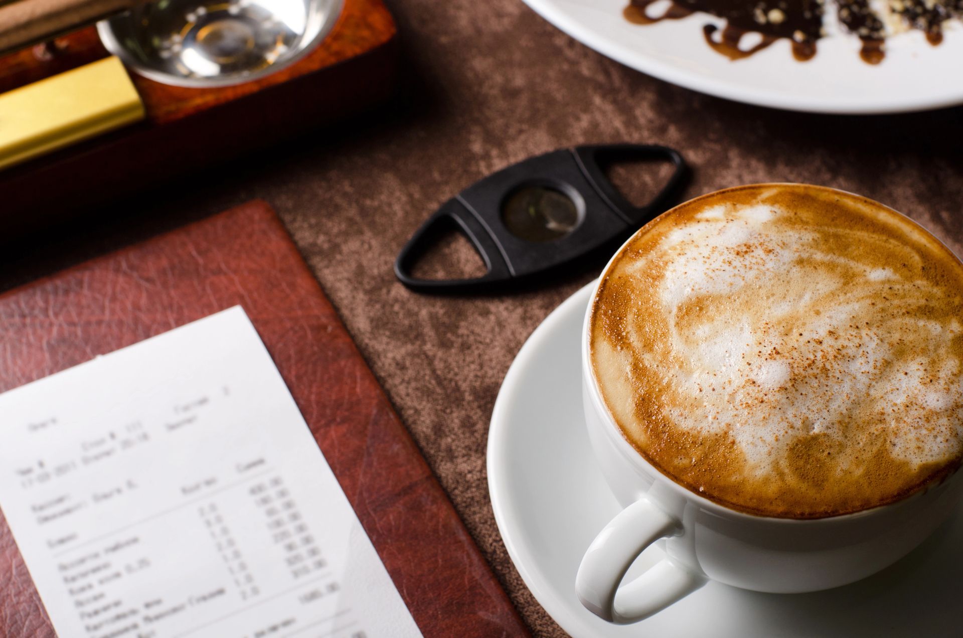 Tazza di cappuccino, scontrino, tagliasigari e parte di un piatto su un tavolo marrone.