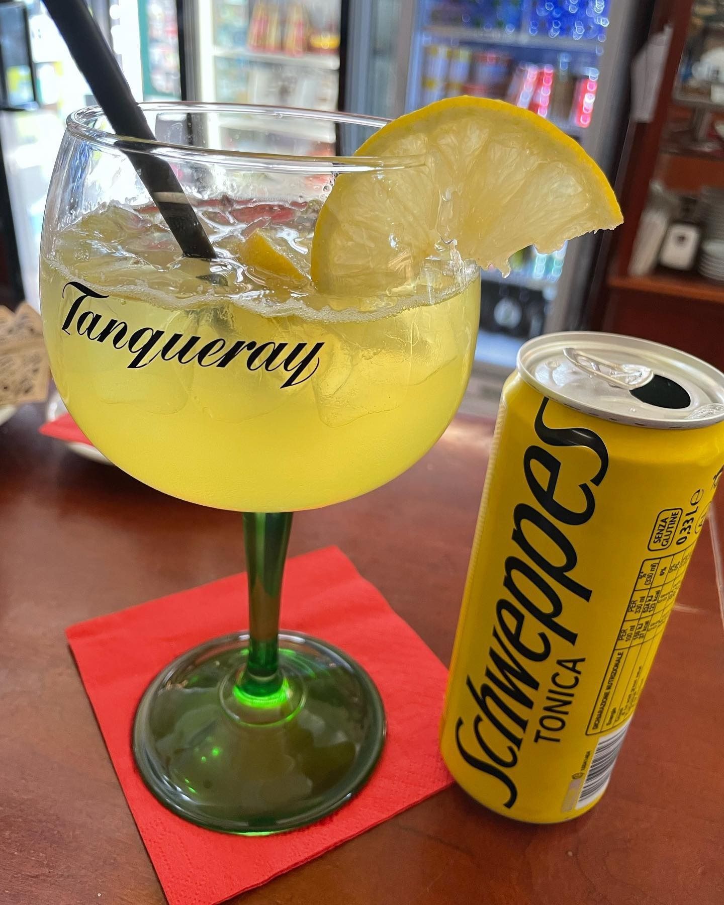 Gin tonic in un bicchiere con stelo, una fetta di limone e una lattina di Schweppes su un tovagliolo rosso.
