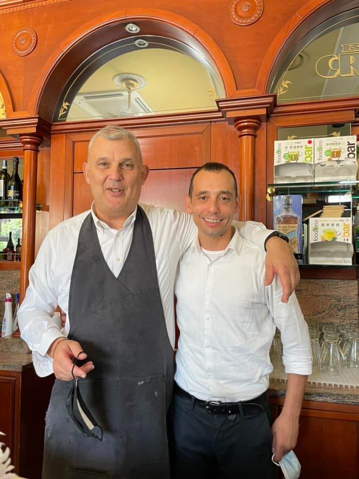 Due uomini sorridenti, uno con il braccio intorno all'altro, in un ristorante arredato con dettagli in legno.