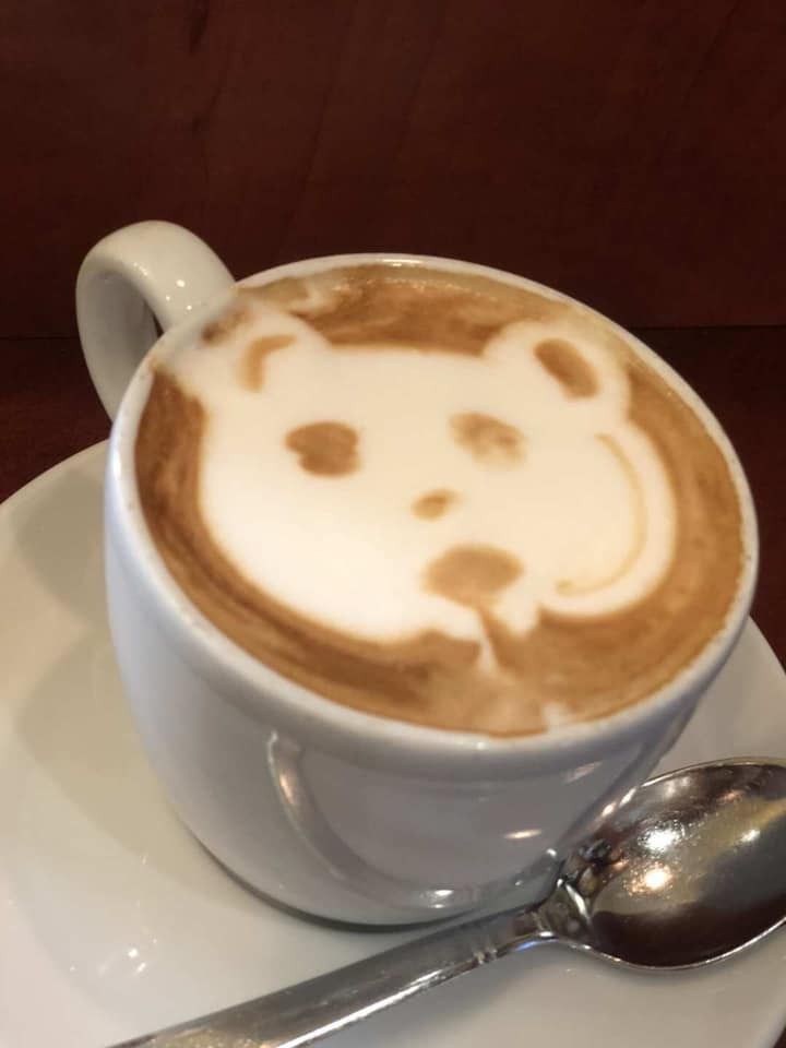 Latte in una tazza bianca, con schiuma di latte raffigurante il muso di un orso; su un piattino con cucchiaio.