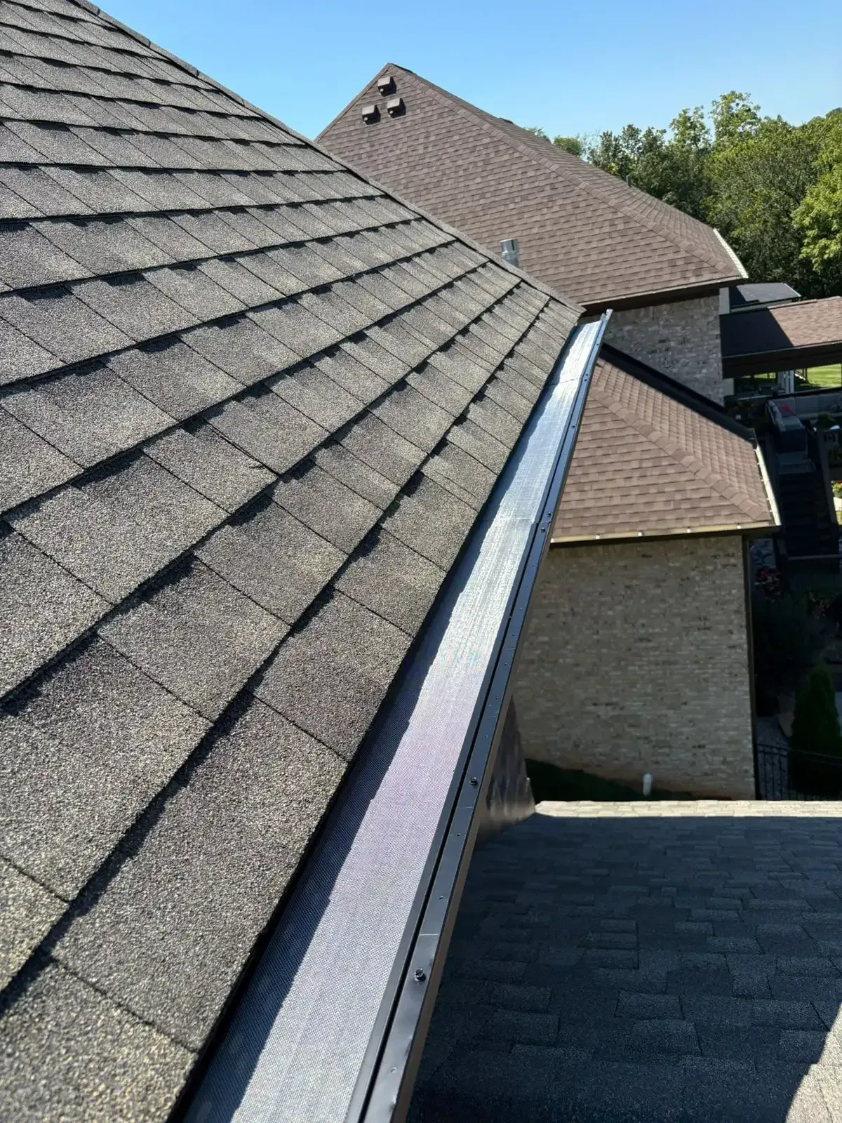 Close-up view of a dark gray shingled roof with a silver metal gutter. The sky is blue.