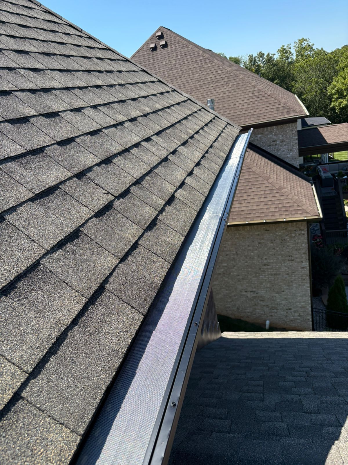 Close-up of a dark asphalt shingle roof with black gutter guards, trees in the background.