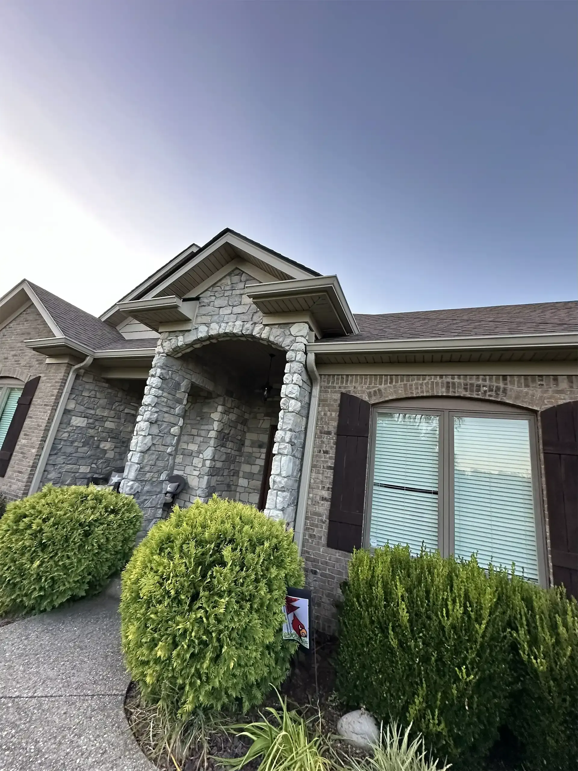 Stone and brick house exterior with a covered entryway and shutters. Green bushes in front.