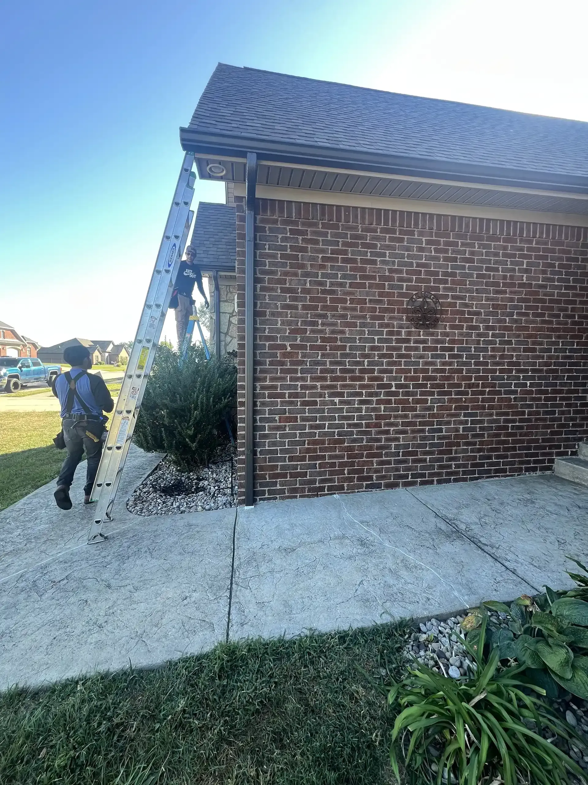 Person on a ladder cleaning gutters on a brick house. Blue sky, greenery, and sidewalk visible.