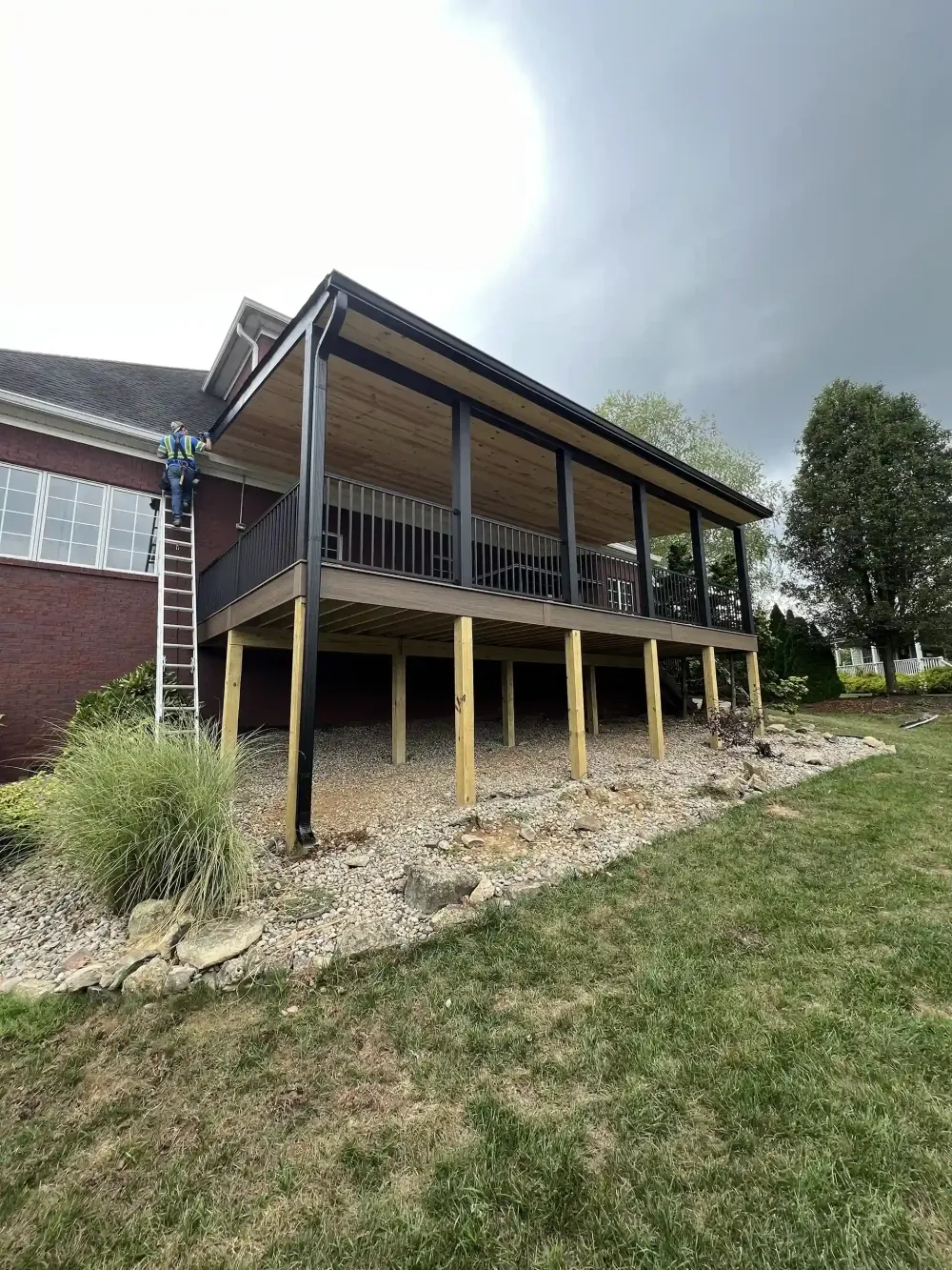 Deck with black railing, brown composite deck, wooden support beams, and a covered roof attached to a brick house.