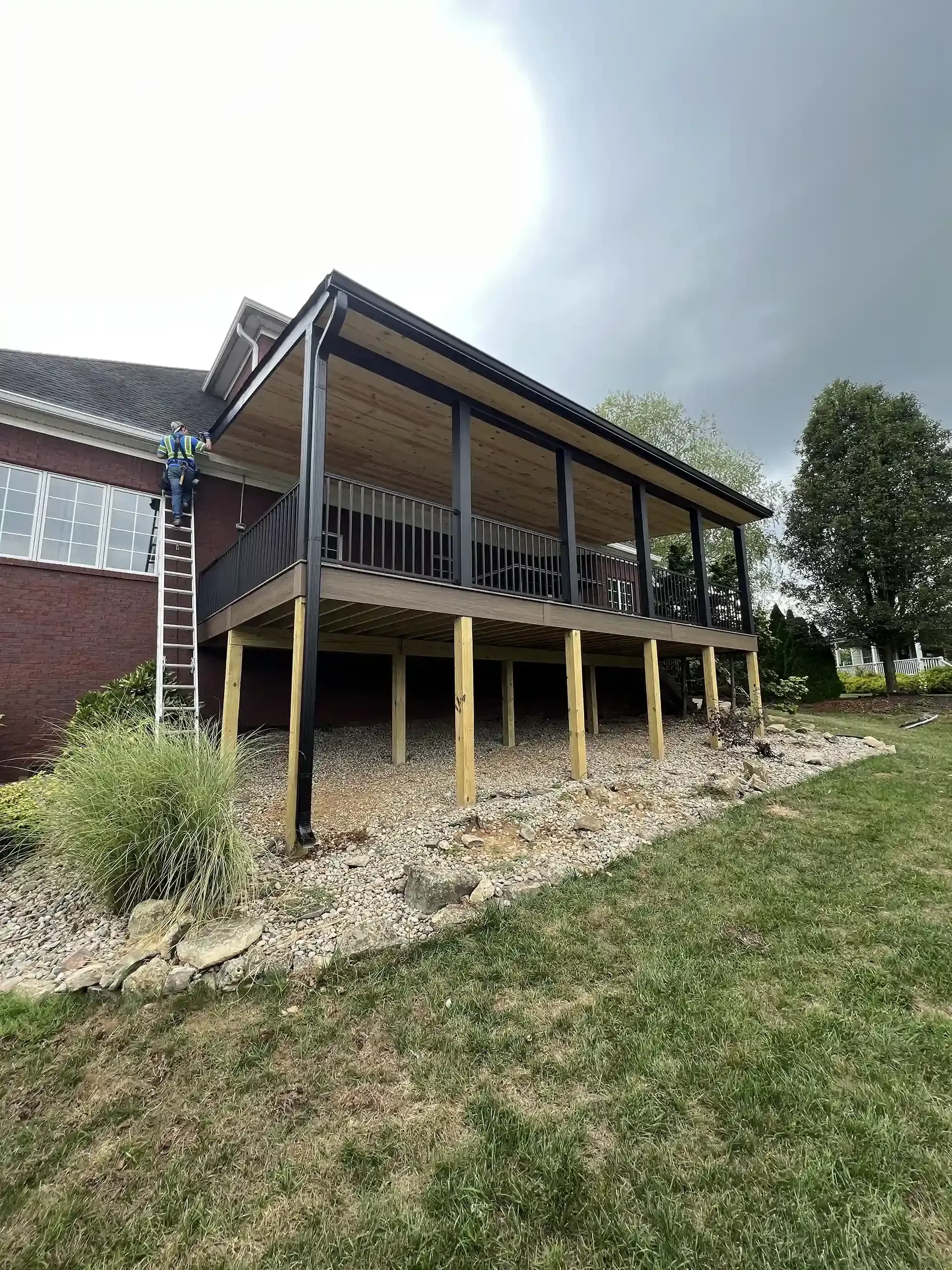 A covered deck attached to a brick house, supported by wooden posts, with a rocky landscaping and overcast sky.