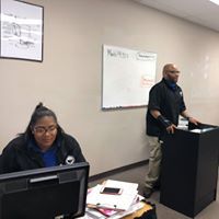 A man is standing at a podium next to a woman sitting at a desk in front of a computer.