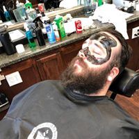 A man with a beard is laying in a barber chair with a black mask on his face.