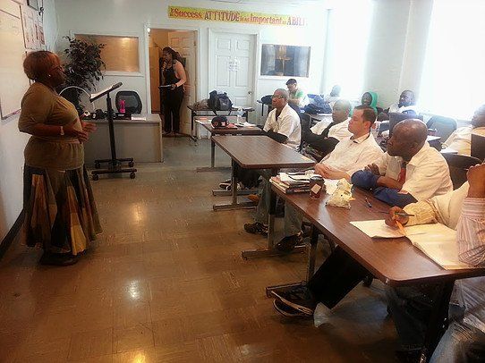 A woman is standing in front of a group of people sitting at tables in a classroom.
