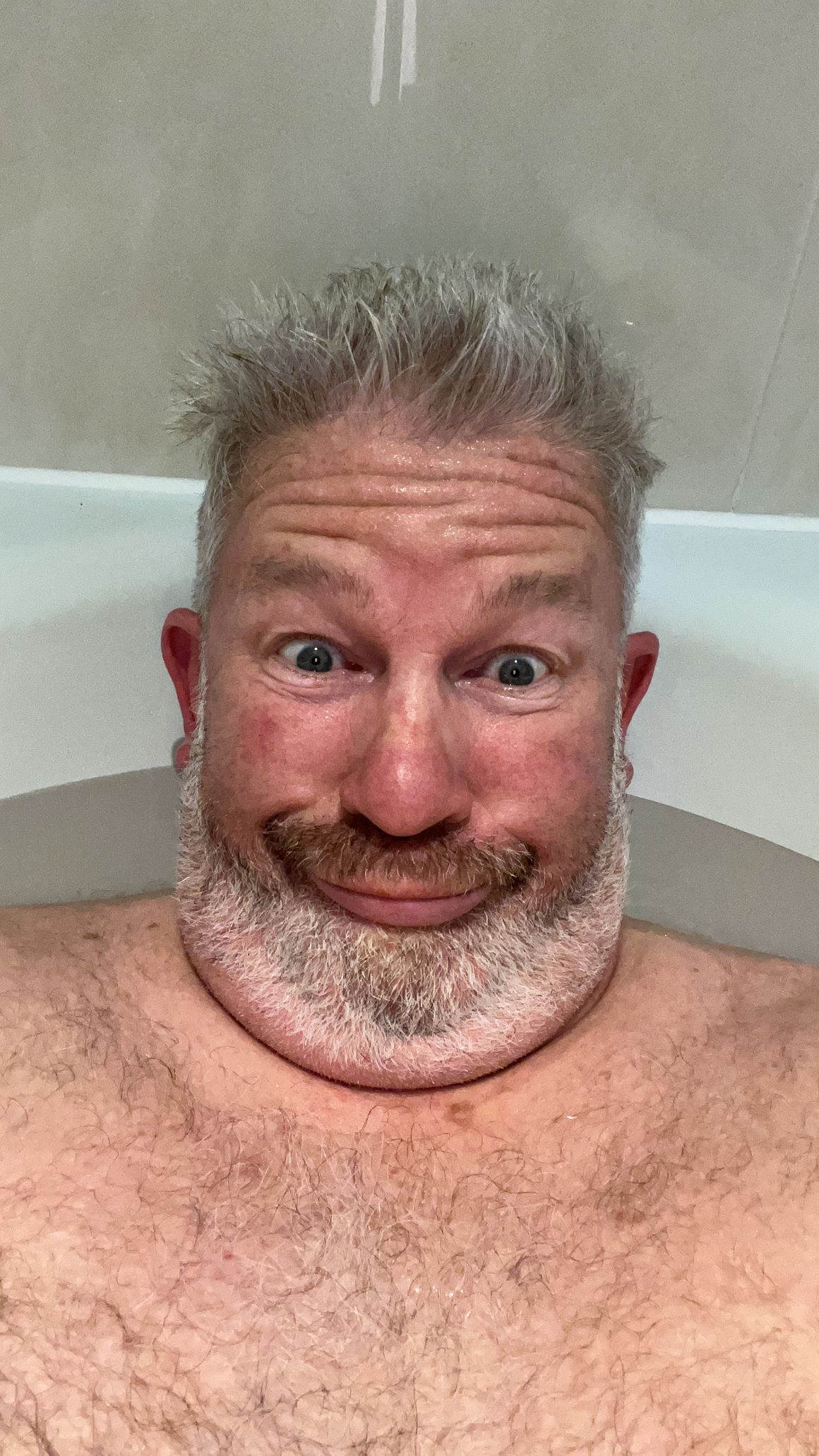Smiling person soaking in a bathtub with a white foam beard and wet spiky hair