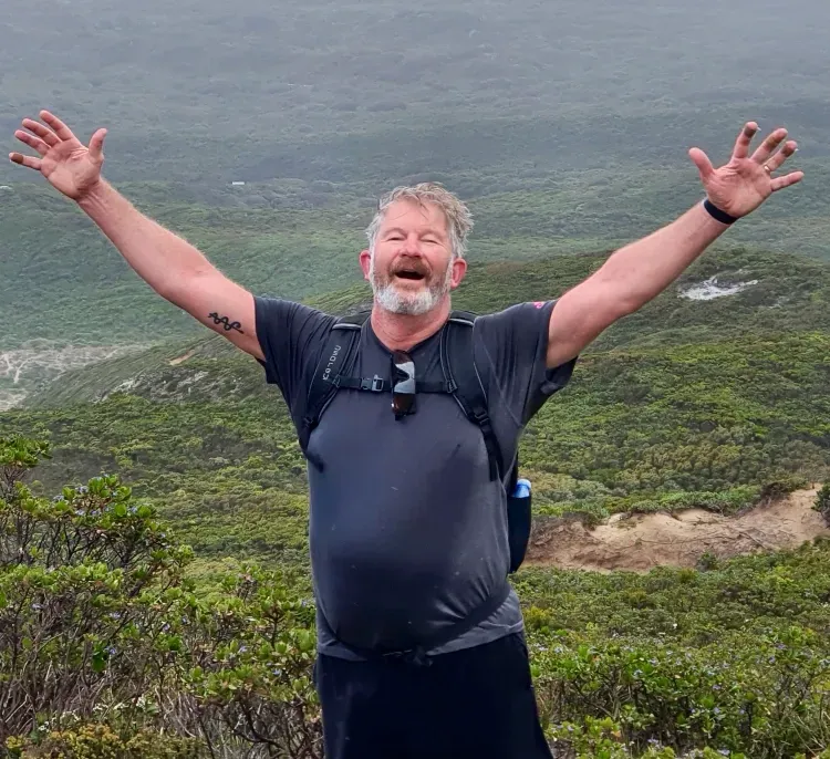 Dr Bruce Powell with his arms outstretched is standing on top of a hill.