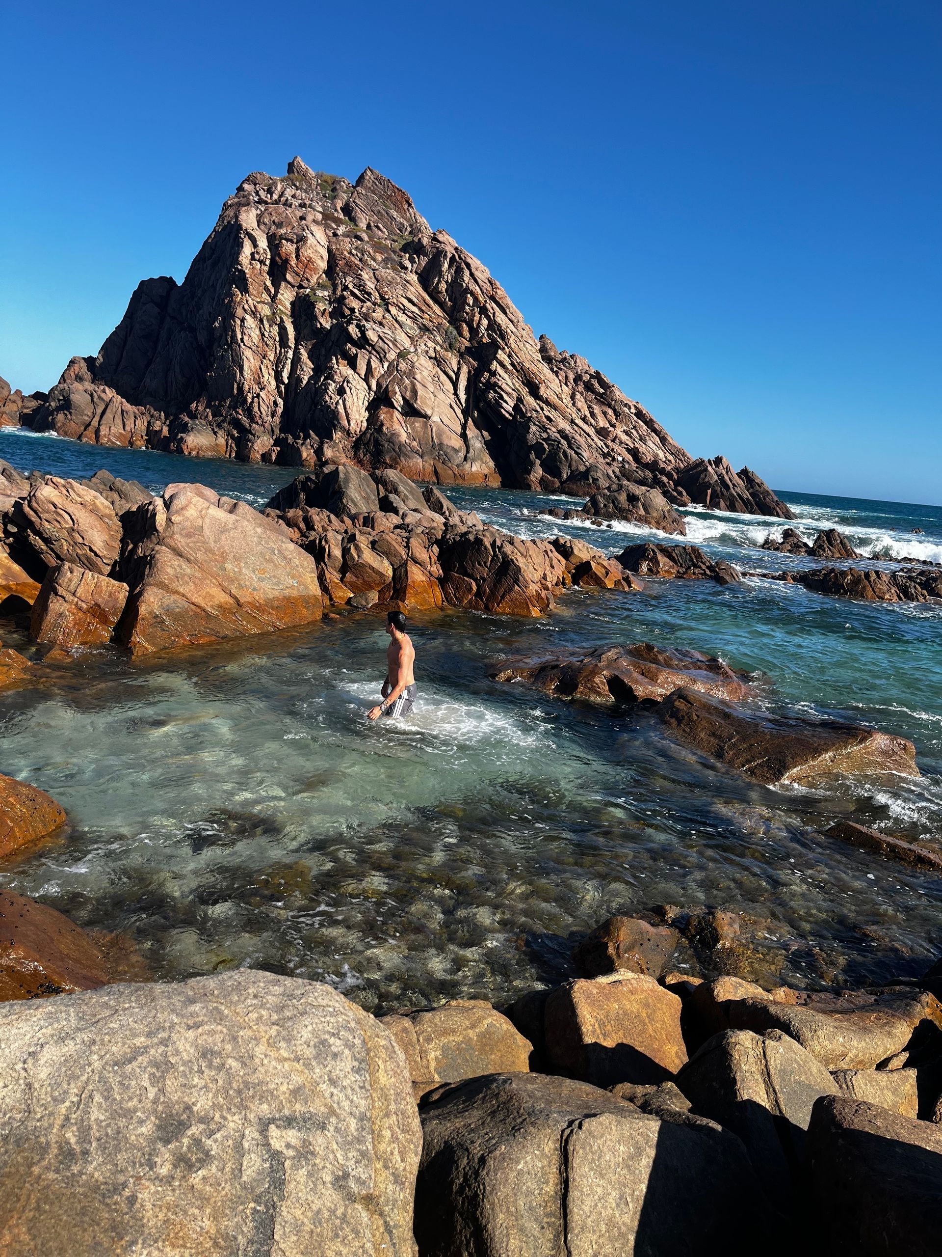 A person wading in a clear, rocky tide pool at the base of a large, craggy mountain under a bright blue sky.