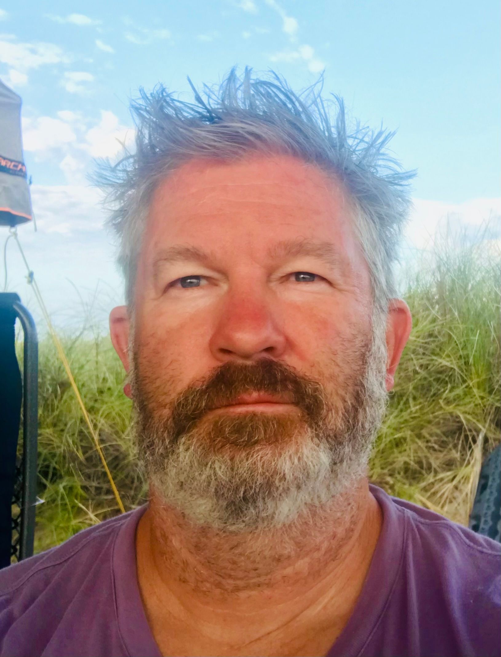 Close-up of a bearded man in a purple shirt outdoors with tall grass and blue sky behind him