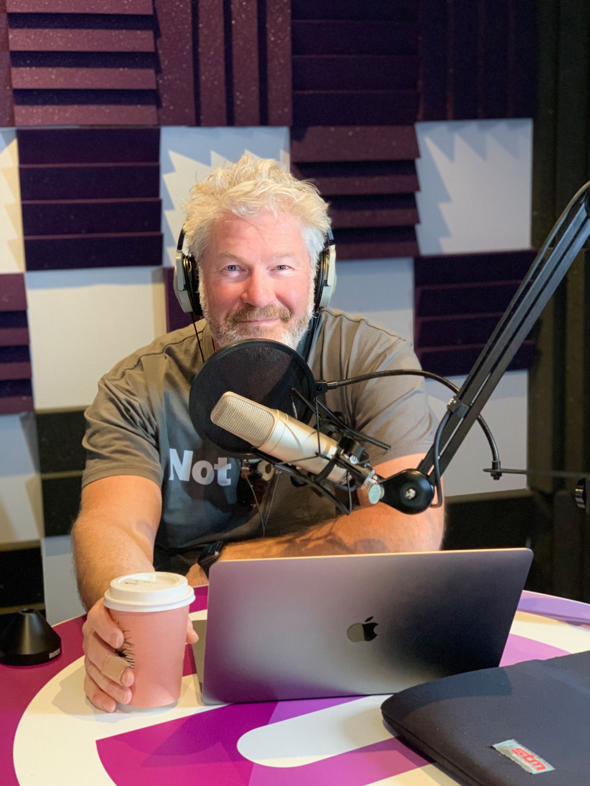 Man at a microphone, in a recording studio, holding a coffee and working on a laptop.