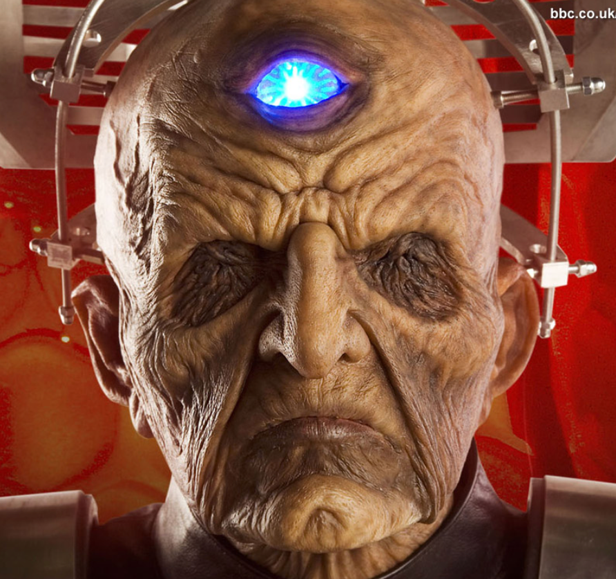 Wrinkled alien face with a glowing blue eye, wearing a metal headpiece.