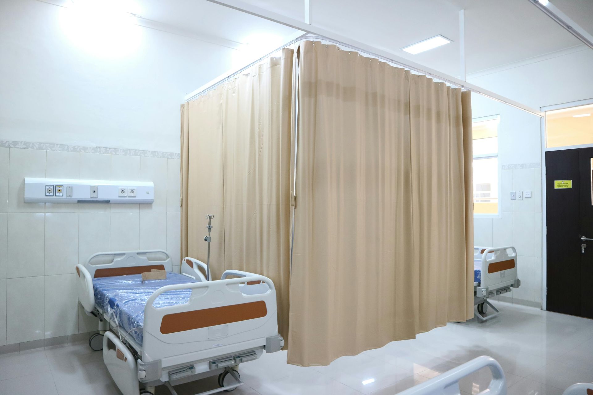 Hospital room with two beds and beige privacy curtains, white walls, and bright overhead lighting