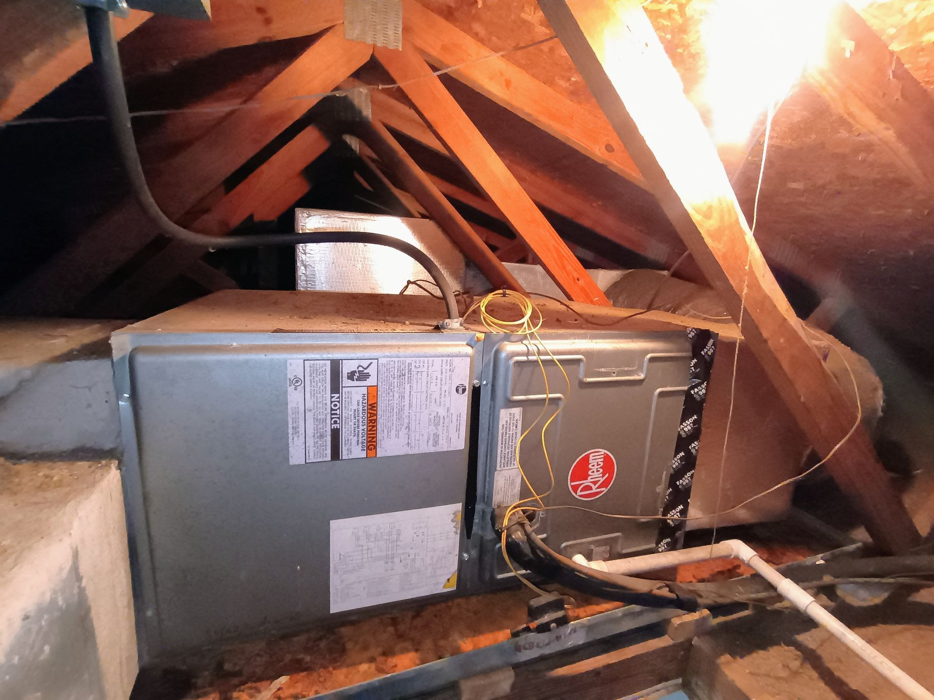 HVAC unit in an attic, gray metal with a Rheem logo, surrounded by wooden beams and insulation.