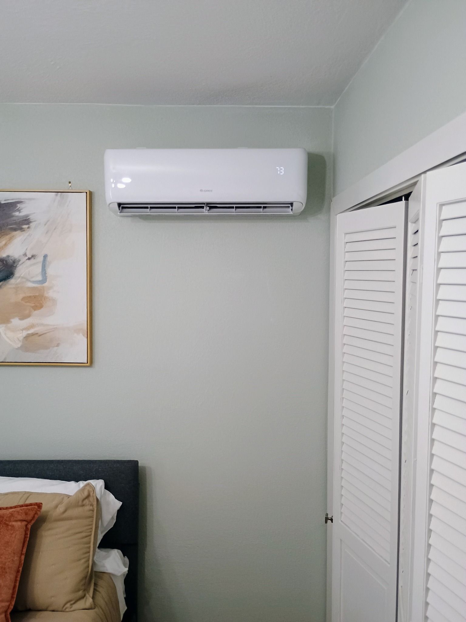 Wall-mounted air conditioner above a bed in a room with a painting and a closet.
