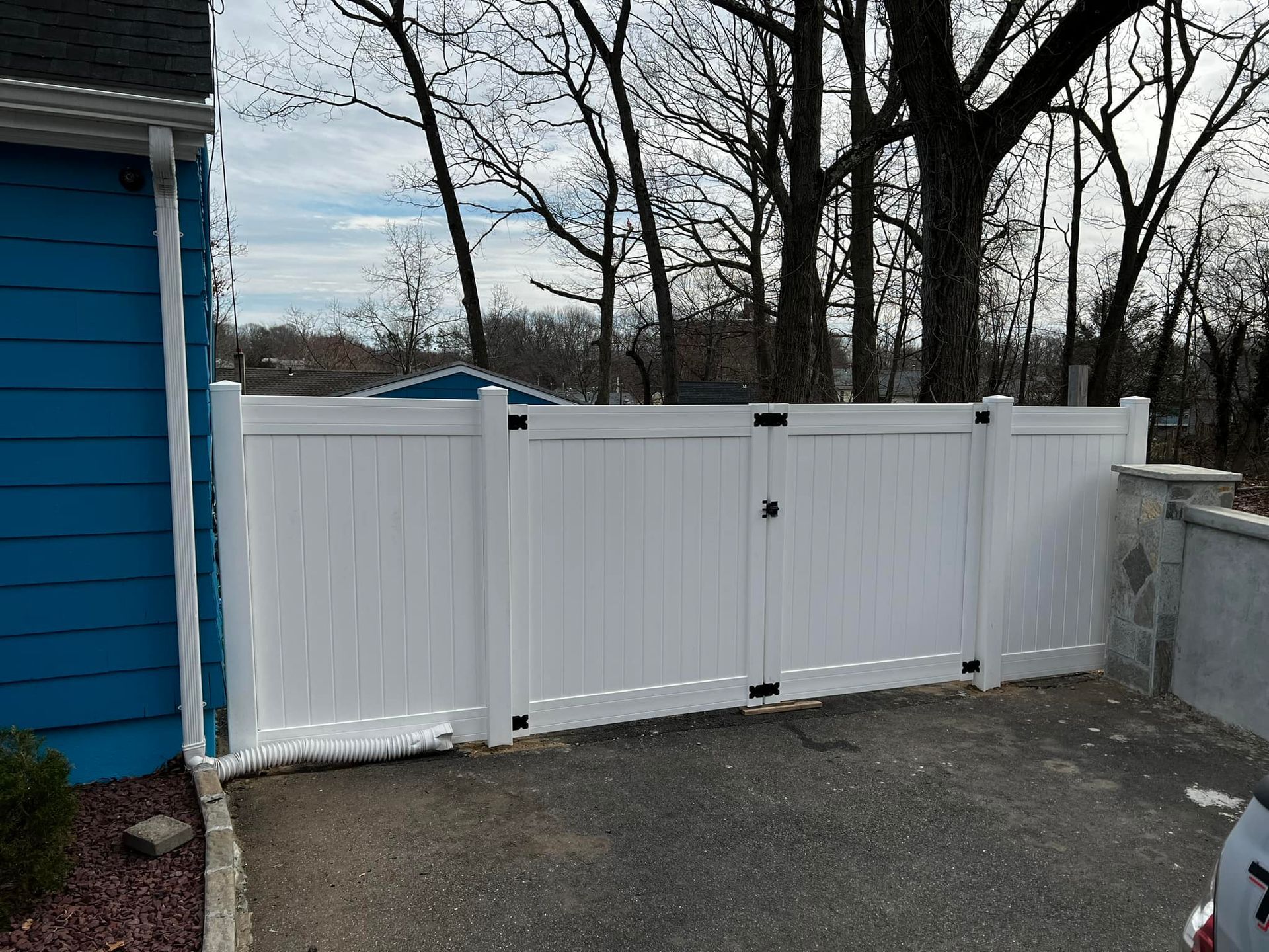 White vinyl gate in front of a blue house and trees. The gate has black hardware and is closed.