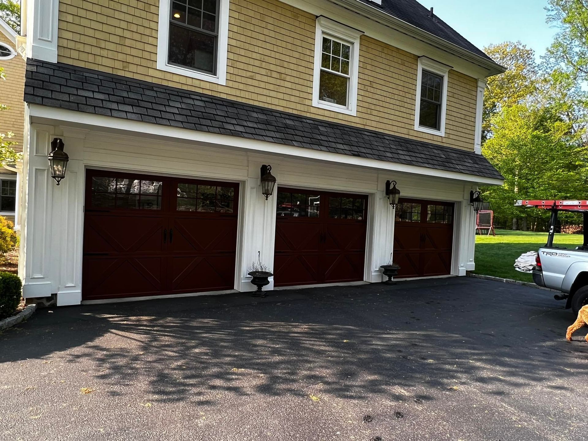 Three-car garage with dark brown doors, white trim, and yellow siding. Black roof and asphalt driveway.