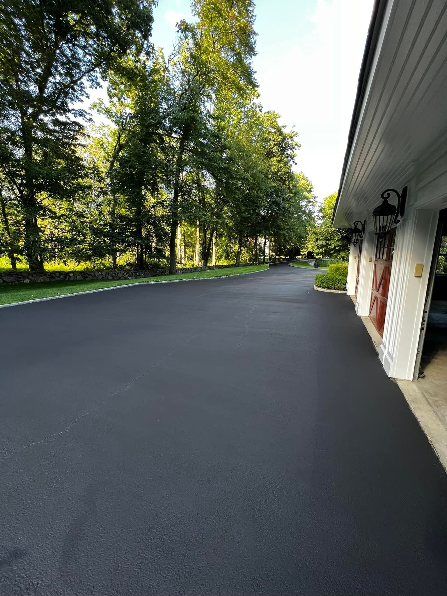 Long, black asphalt driveway next to a white garage, leading into a wooded area.