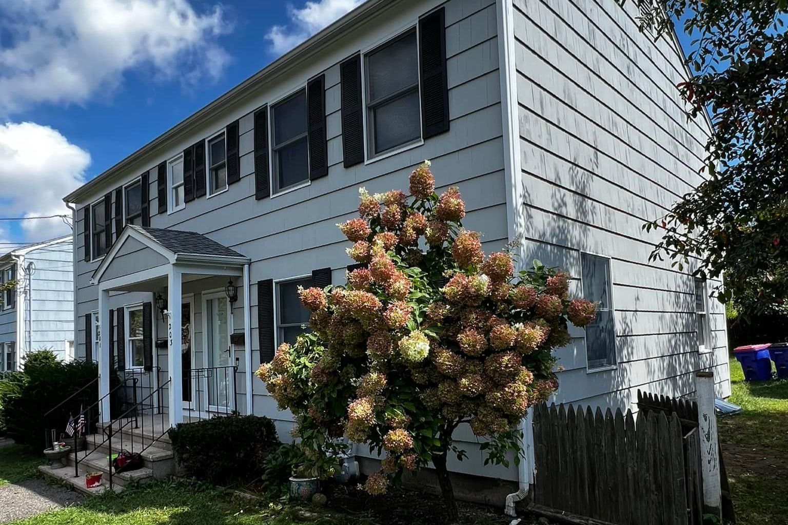 Two-story gray building with black shutters, white trim, and a porch. A large bush in the foreground.