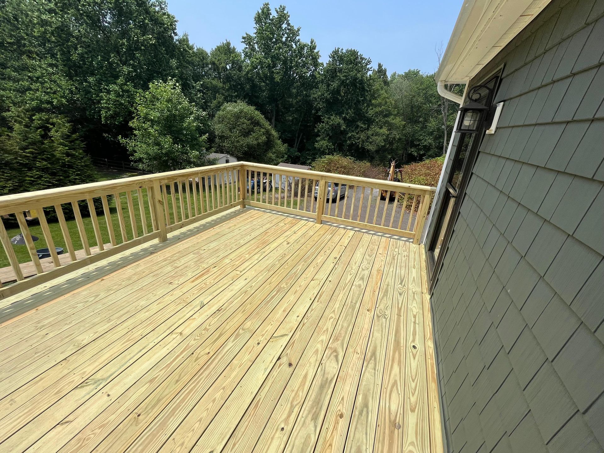 Wooden deck with railing, attached to a house with green siding, overlooking trees and a yard on a sunny day.