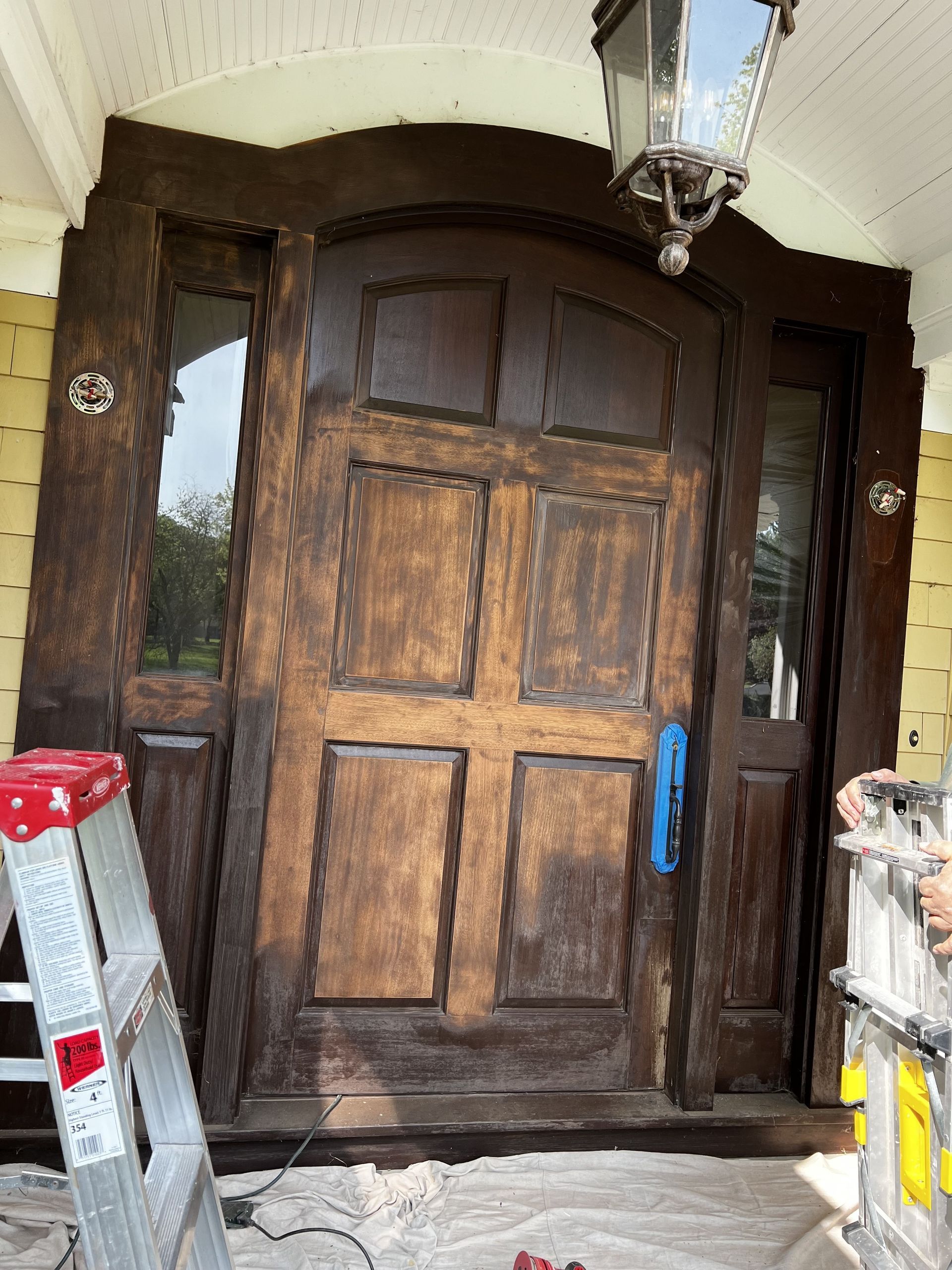 Wooden front door with sidelights, under a porch overhang, being worked on.