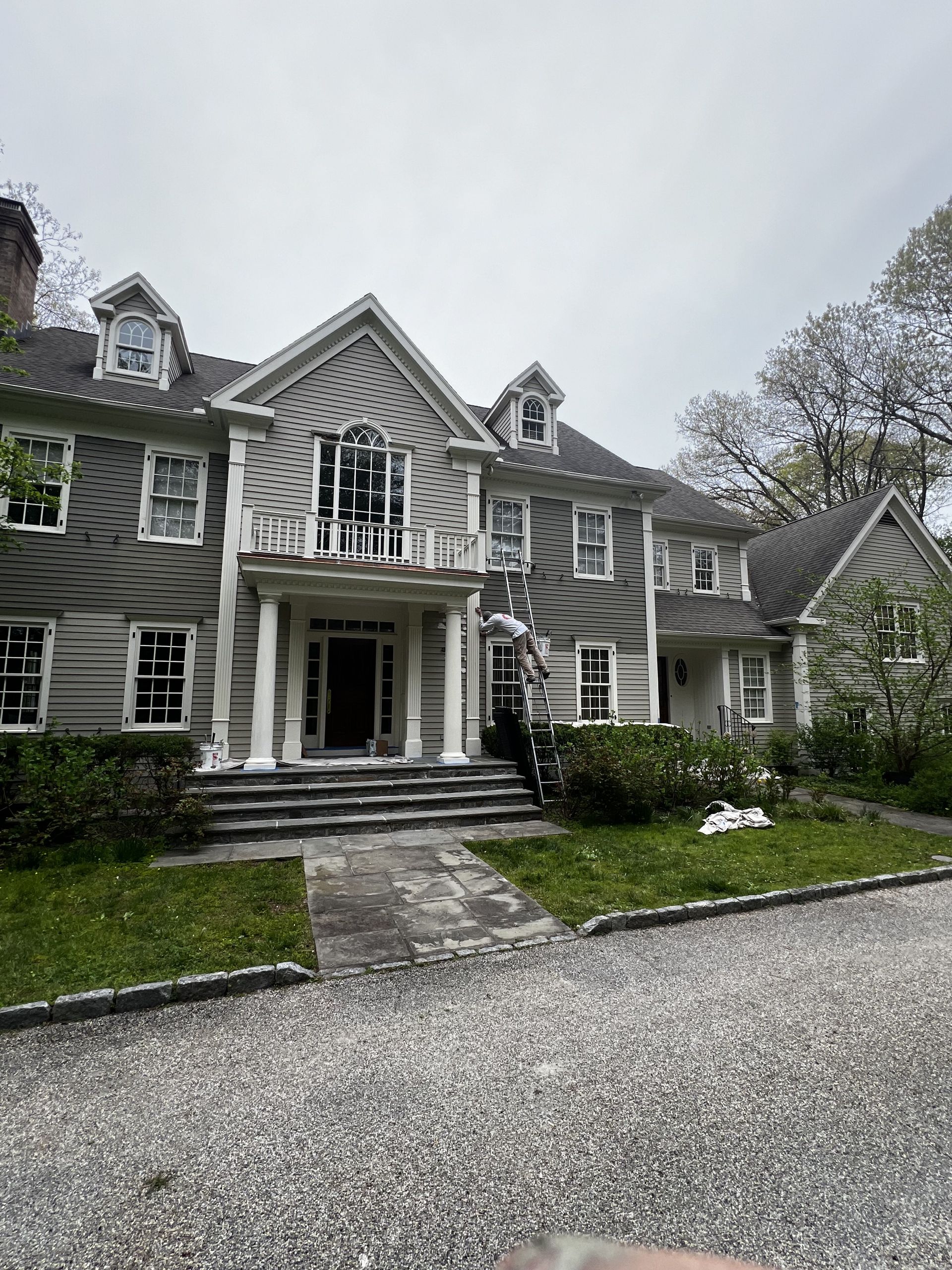 Large gray two-story house with white columns, balcony, and dormer windows. Gravel driveway, green grass, overcast sky.
