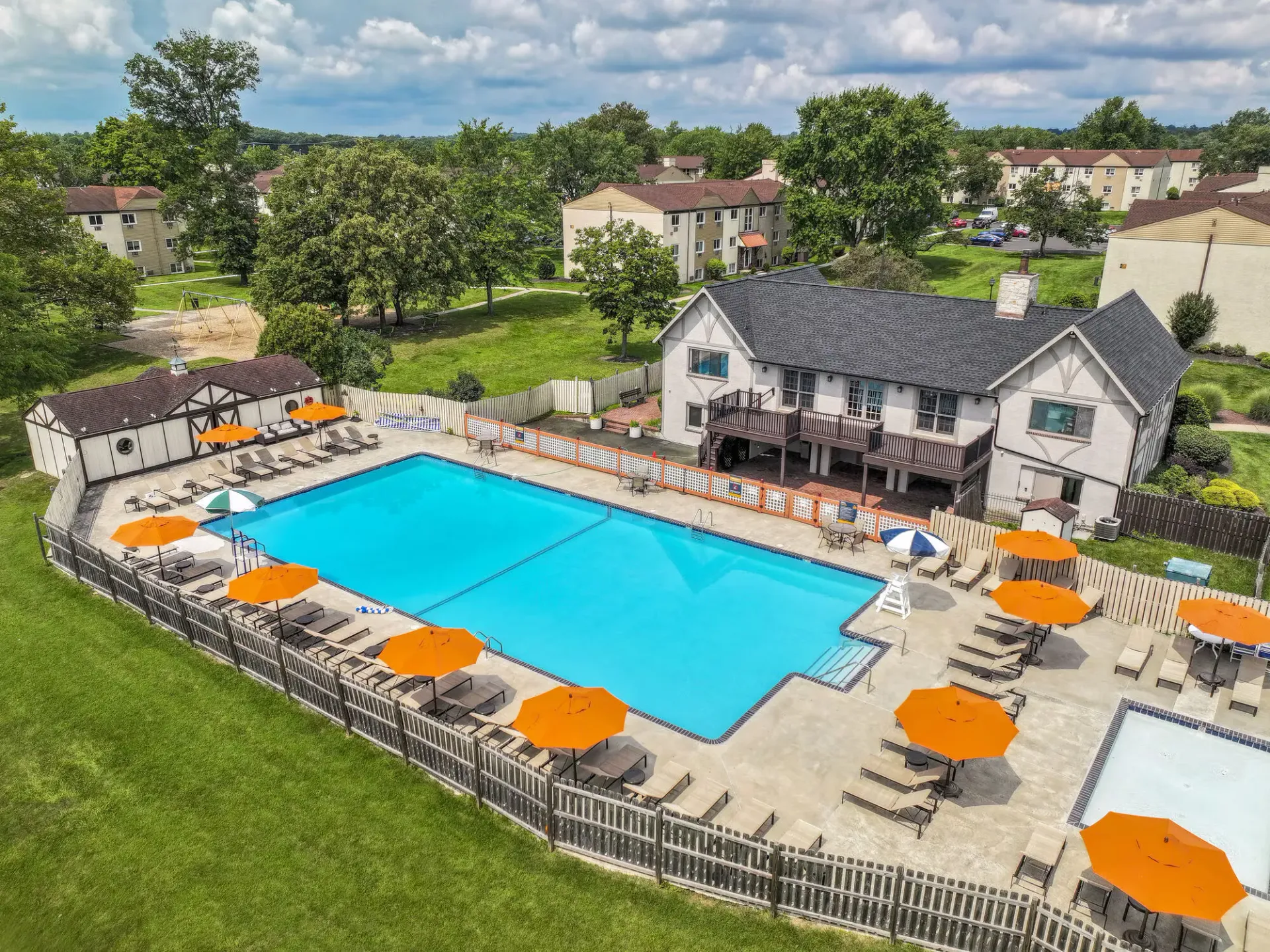 Aerial view of a community pool area with orange umbrellas and lounge chairs beside a clubhouse at The Addison, offers apartments in North Wales, PA.