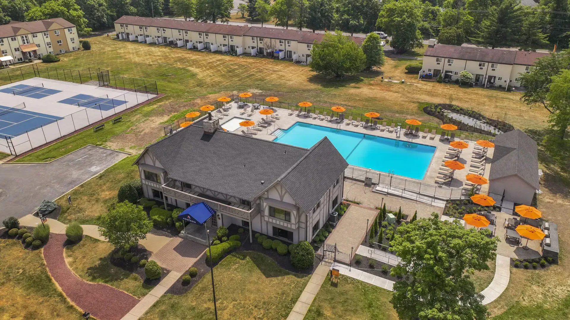 Aerial view of a residential pool area with orange umbrellas, lounge chairs, and a clubhouse at The Addison, offers apartments for rent in North Wales, PA.