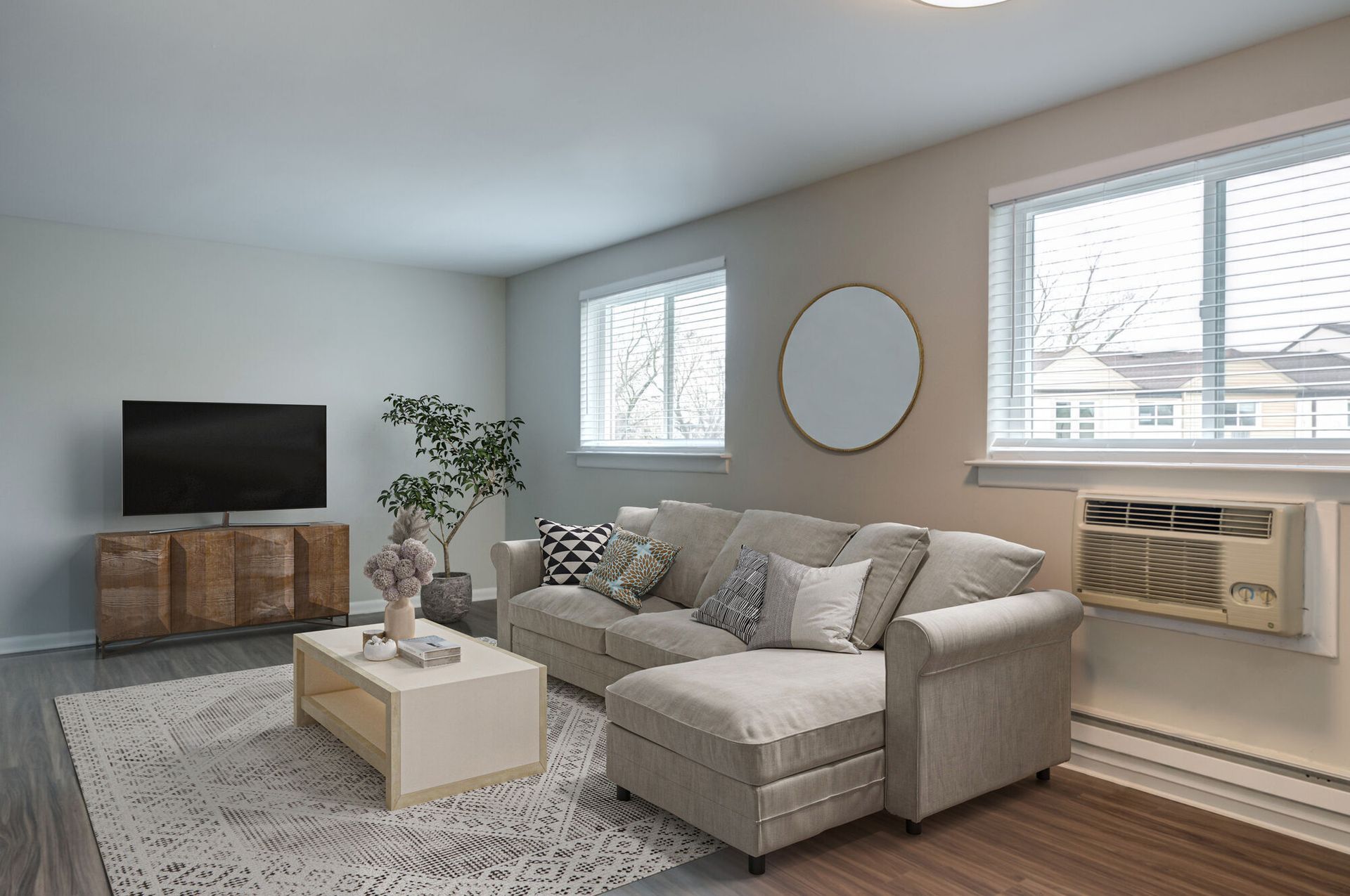 Living room with a gray sectional sofa, coffee table, rug, TV on a wooden console, and a window AC unit at The Addison, offers townhomes in North Wales, PA.