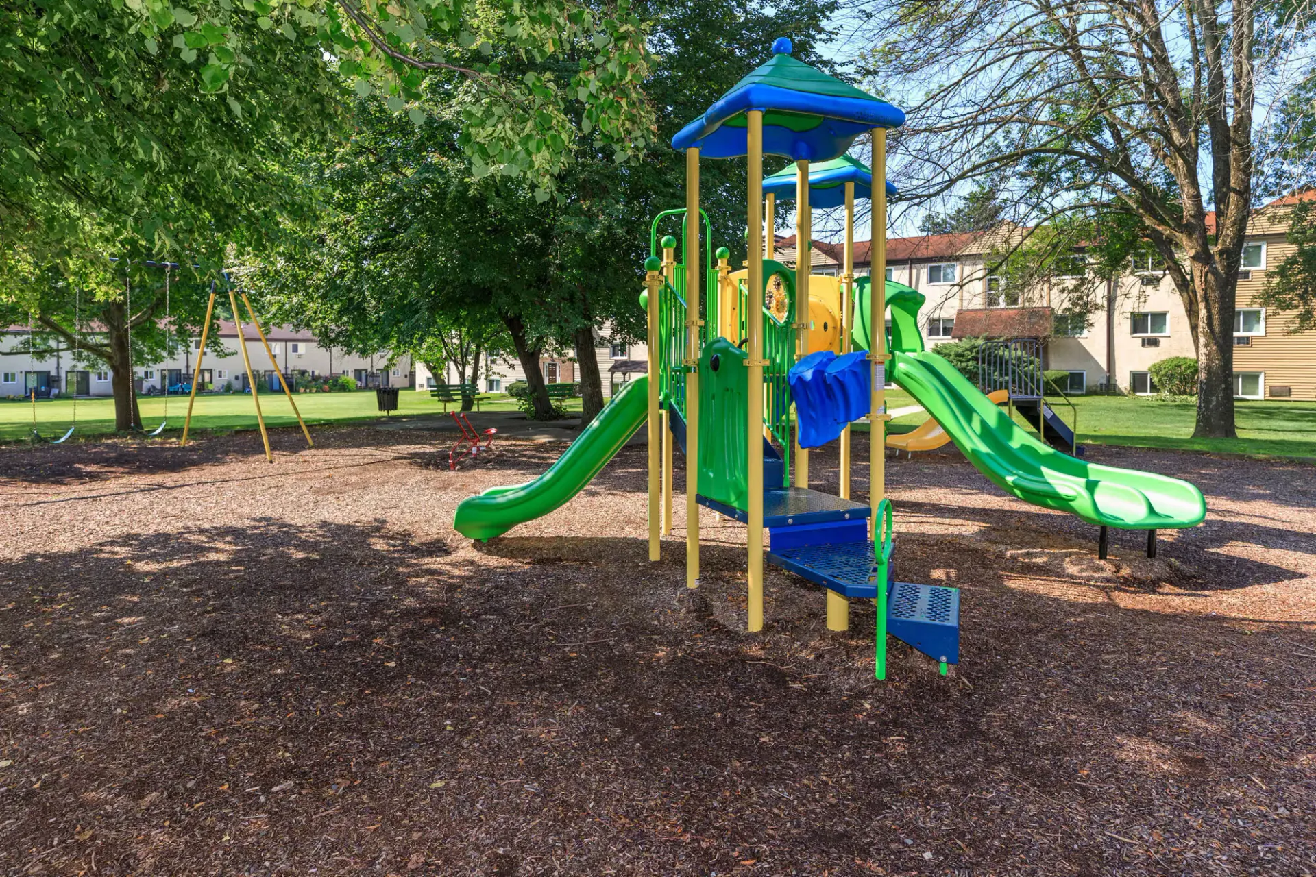 Colorful playground with slides and swings under trees in a residential courtyard at The Addison, offers apartments in North Wales, PA.