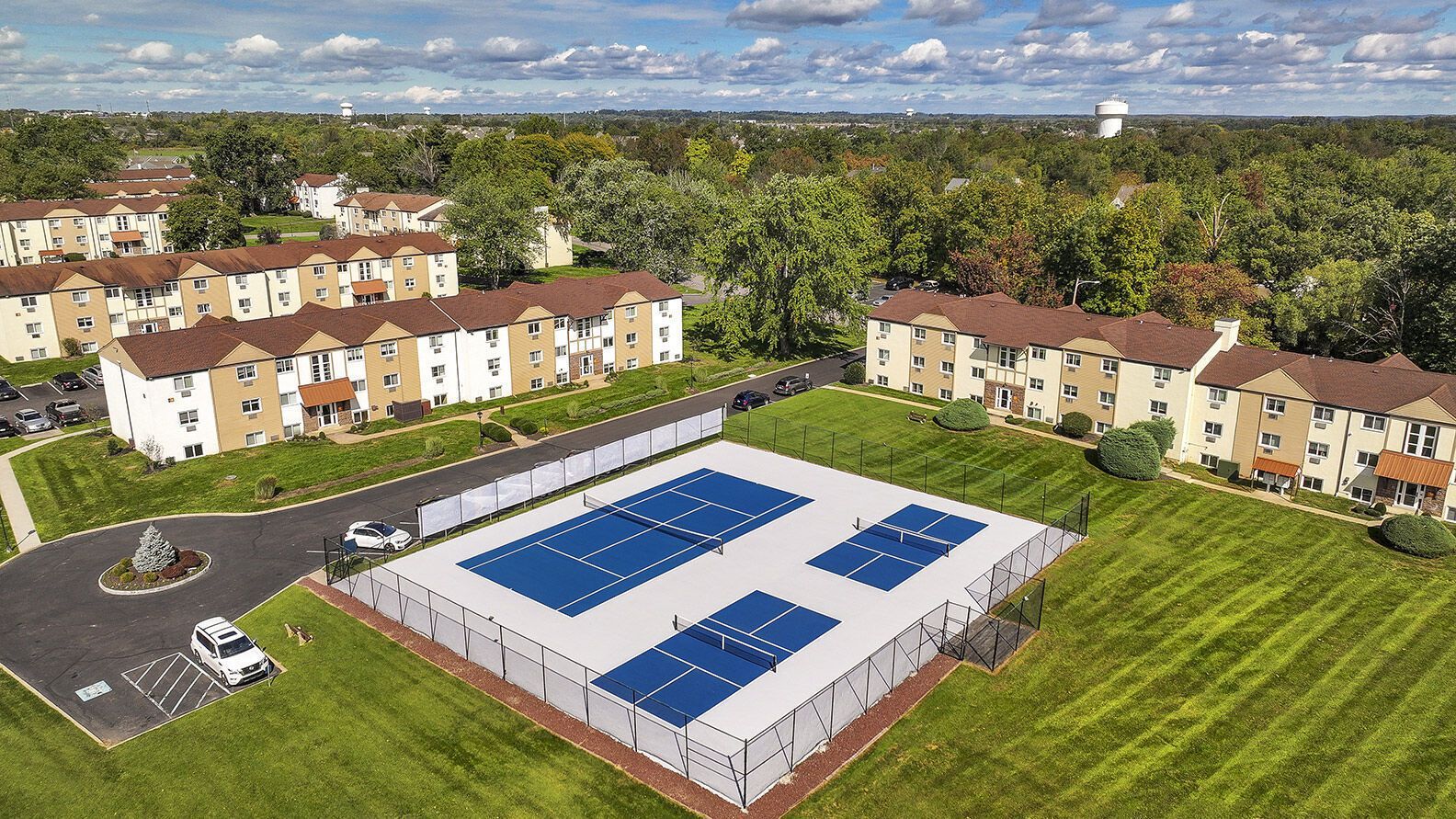 Aerial view of a beige apartment complex with a fenced blue tennis/pickleball court at the center at The Addison, offers apartments for rent in North Wales, PA.