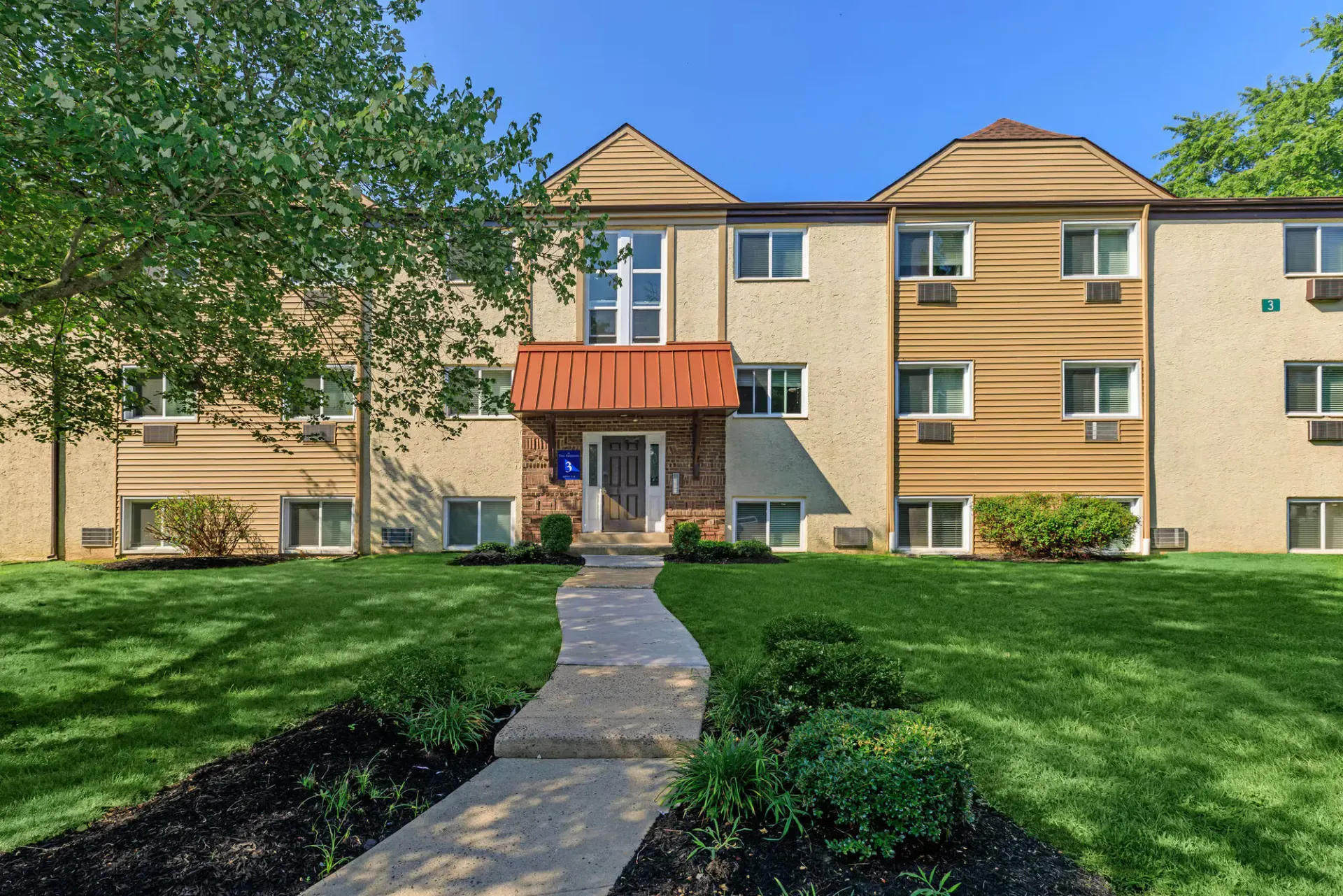 Pathway to central building entrance with a small stoop, brick façade, and well-kept lawn at The Addison, offers apartments for rent in North Wales, PA.