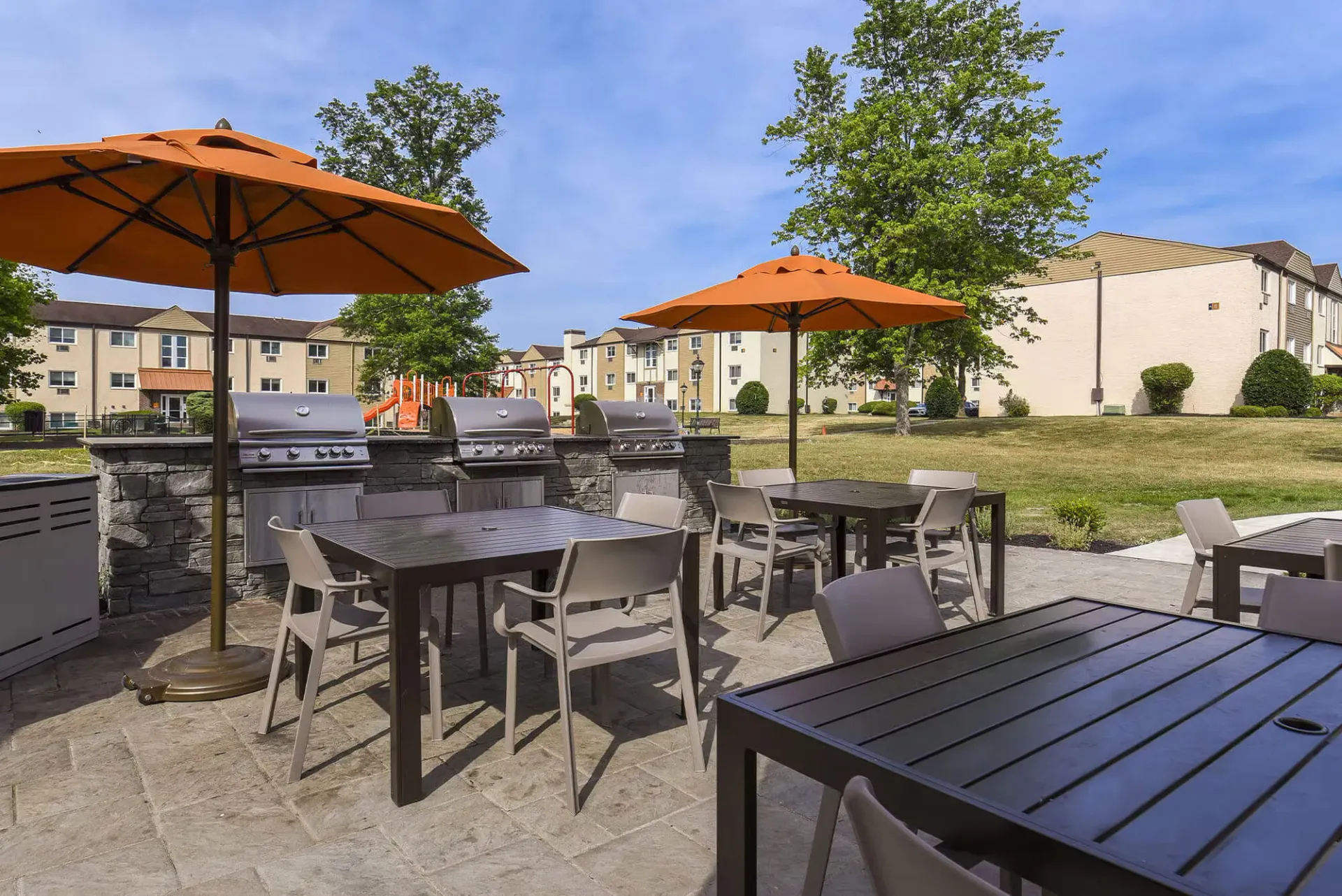 Outdoor communal grilling area with tables under orange umbrellas, playground in the background at The Addison, offers apartments for rent in North Wales, PA.
