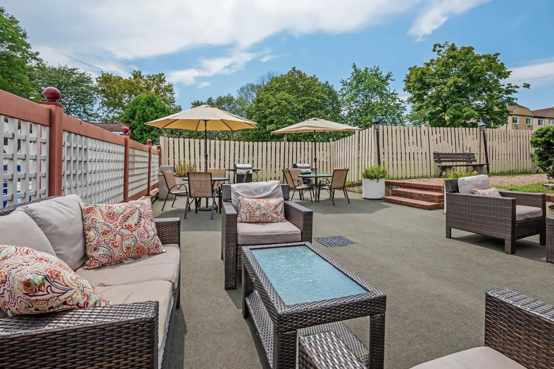 Outdoor communal patio with wicker seating, a glass-top coffee table, umbrellas, and a wooden privacy fence at The Addison, offers apartments for rent in North Wales, PA.