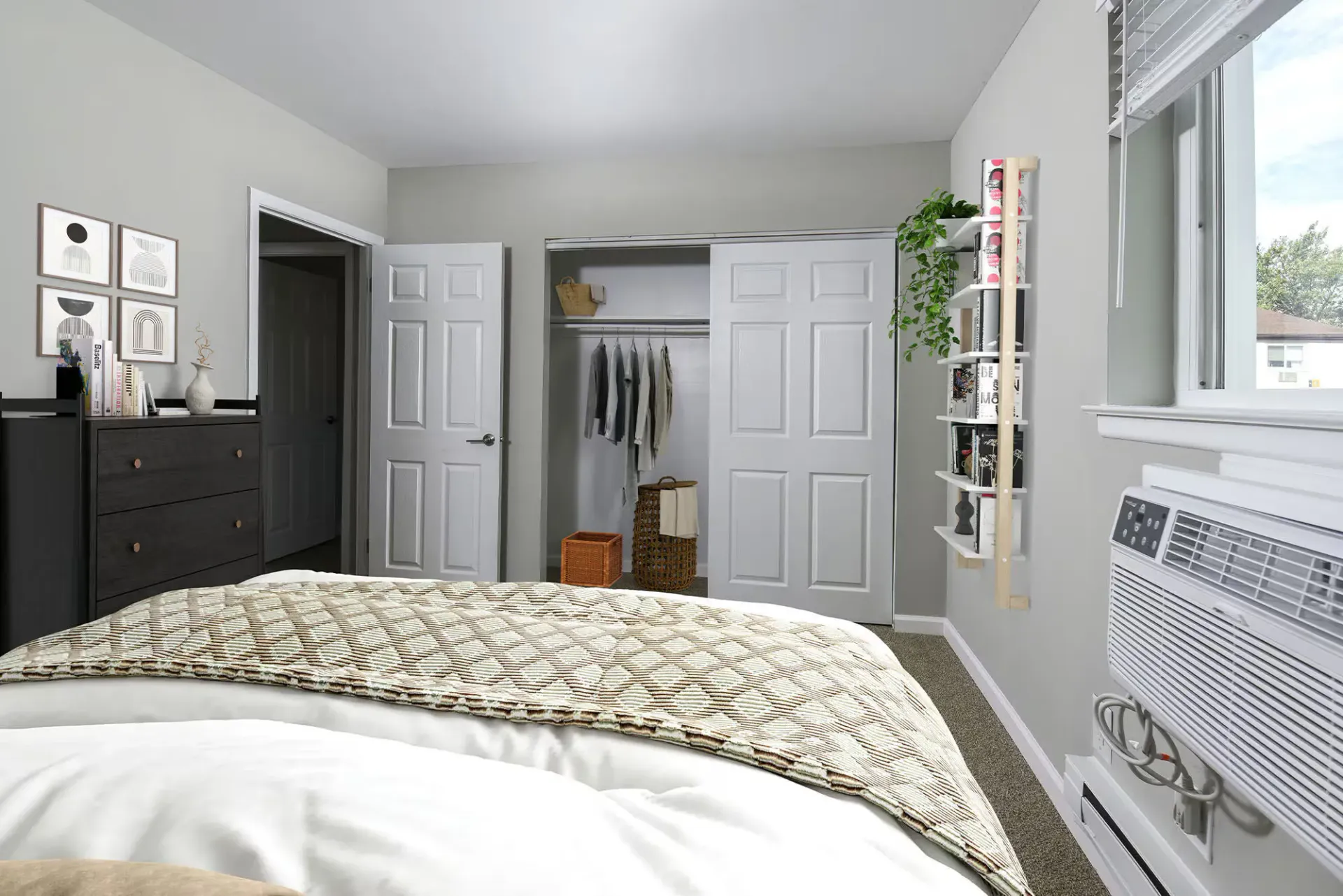 Bedroom interior with bed, dresser, open closet, window air conditioner, and shelving at The Addison, offers townhomes for rent in North Wales near Delaware Valley University.