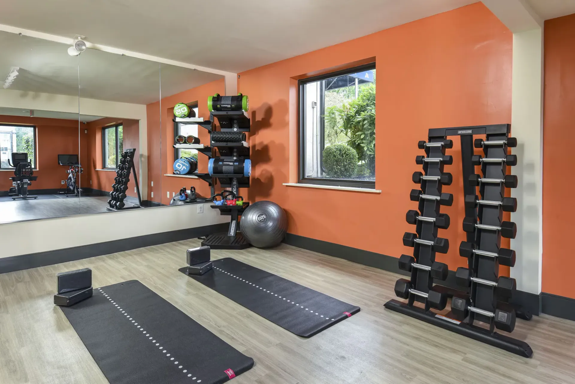 Indoor community gym with orange walls, mirrors, dumbbells, and exercise mats at The Addison, offers apartments in North Wales, PA.
