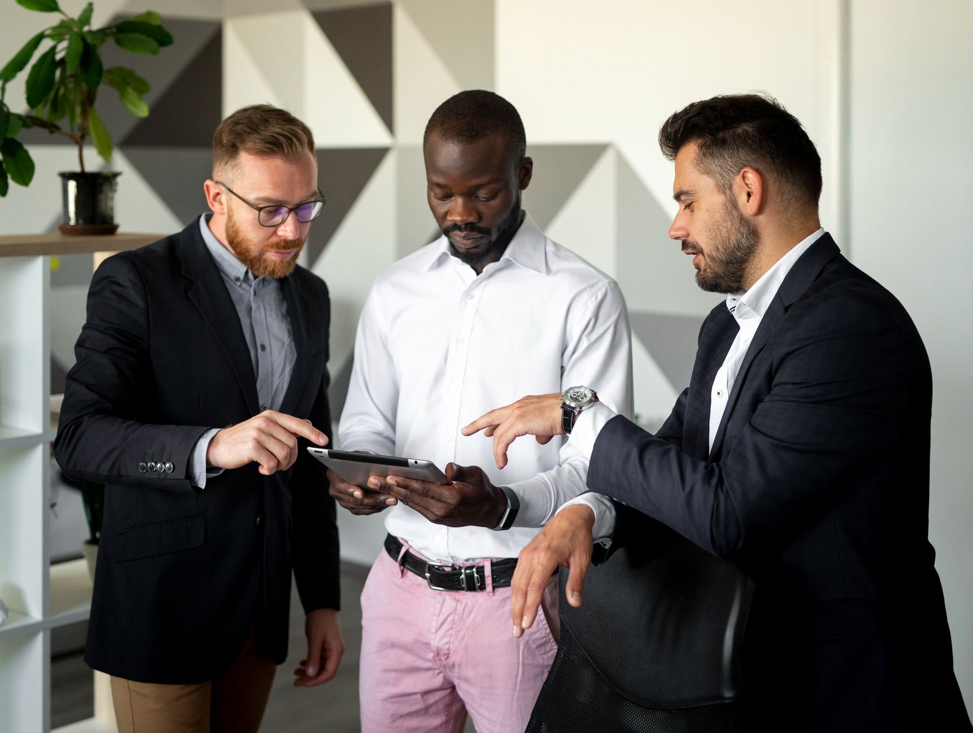 Three men in business attire looking at a tablet in an office setting. The man on the left points at the screen.