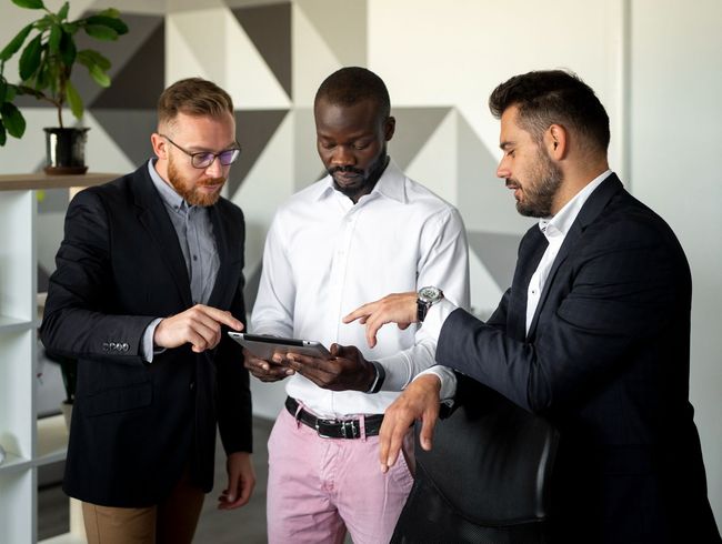 Three men in business attire looking at a tablet in an office setting. The man on the left points at the screen.